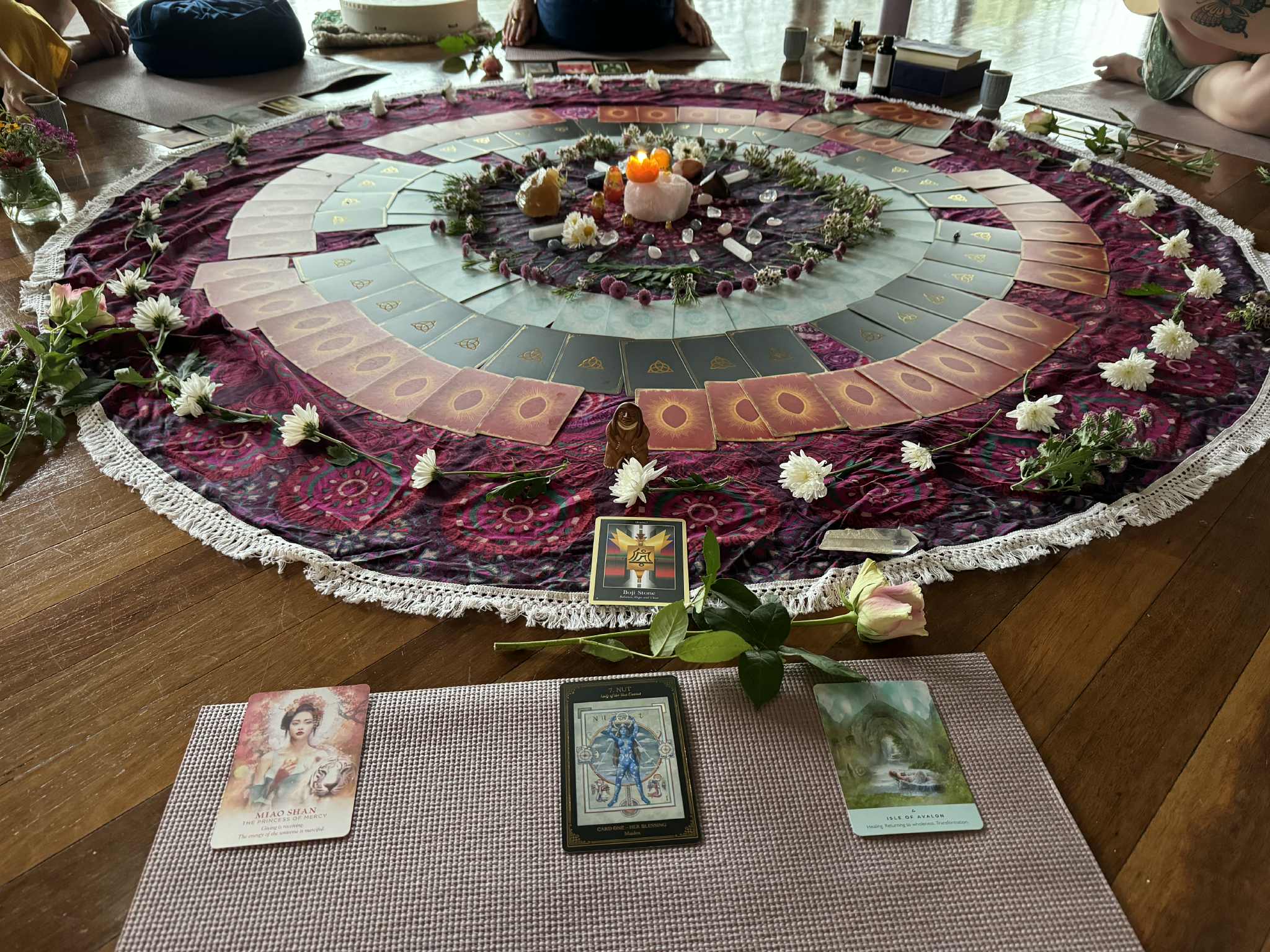 A beautiful altar with concentric rings of oracle cards, crystals, a candle, and white flowers on a purple cloth
